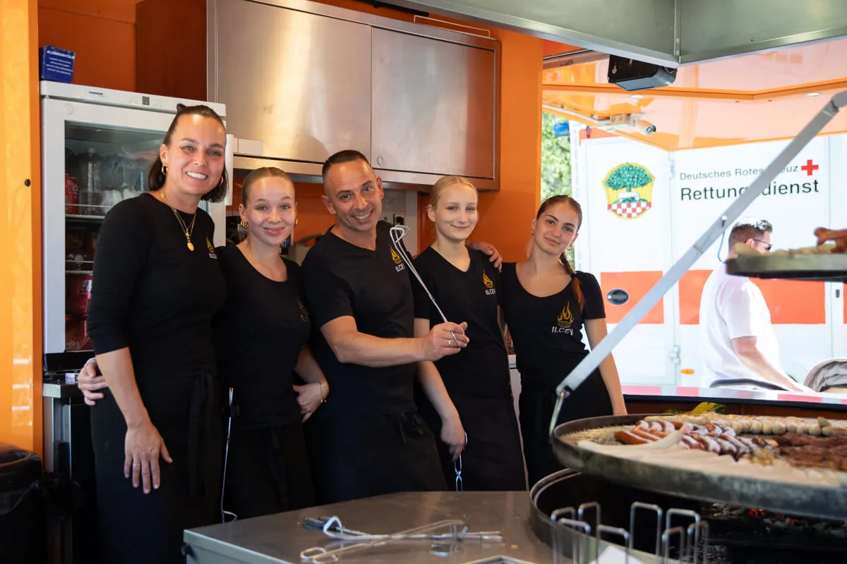 Fünf Personen in schwarzer Arbeitskleidung stehen vor einem orangefarbenen Imbisswagen mit dem Logo des Deutschen Roten Kreuzes und bereiten Speisen zu.