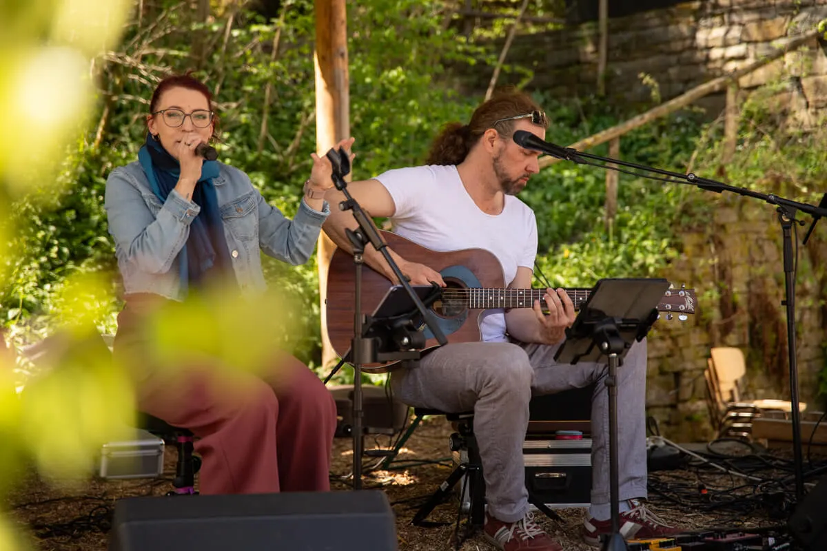 Duo spielt akustische Musik im Freien. Eine Frau singt in ein Mikrofon, während ein Mann Gitarre spielt. Im Hintergrund grüne Bäume und Büsche.