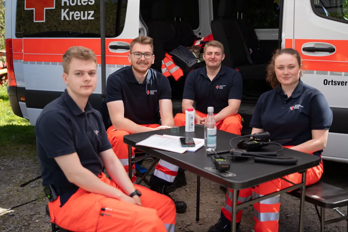 Vier junge Einsatzkräfte des Deutschen Roten Kreuzes sitzen vor einem Behandlungstisch und einem Krankenwagen. Sie tragen dunkelblaue Poloshirts mit Logo und orangefarbene Hosen.