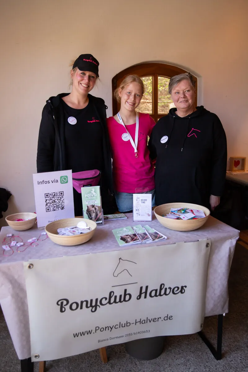Drei Frauen stehen hinter einem Tisch mit Informationsmaterial des Ponyclubs Halver, inklusive QR-Code und Kontaktinformationen. Im Hintergrund ist ein Gebäude mit einem Fenster zu sehen.