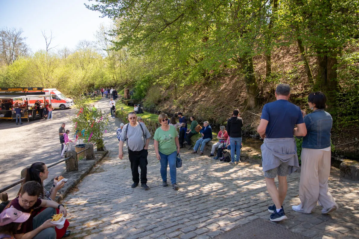Menschen flanieren und unterhalten sich auf einem gepflasterten Weg vor der Heesfelder Mühle, während im Hintergrund ein Marktstand und ein rotes Fahrzeug zu sehen sind.
