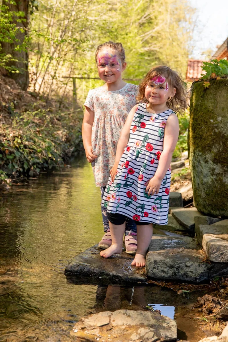 Zwei lächelnde Mädchen mit Gesichtsbemalung stehen an einem Bachlauf, die Füße im Wasser. Im Hintergrund eine grüne Landschaft und ein Steinbau.