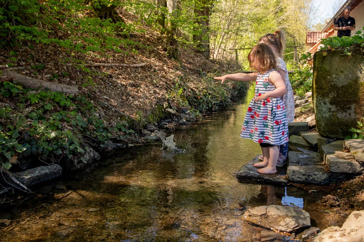 Ein kleines Mädchen steht barfuß auf Steinen in einem Bach und berührt das Wasser. Im Hintergrund eine Frau und ein bewaldeter Hang.