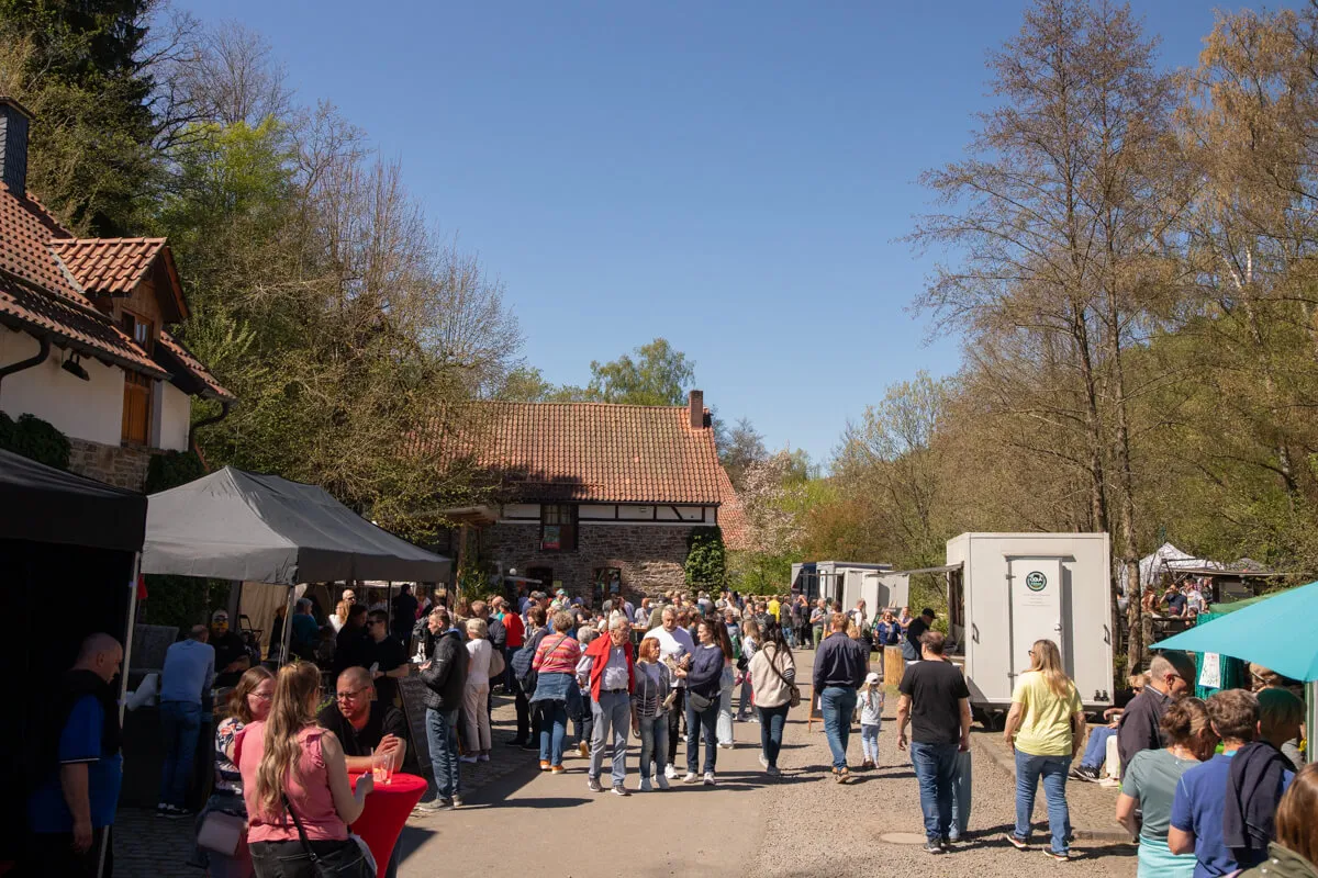 Menschenmengen bewegen sich auf einem belebten Marktplatz vor historischen Fachwerkhäusern mit roten Ziegeldächern. Im Hintergrund sind Bäume und ein blauer Himmel zu sehen.