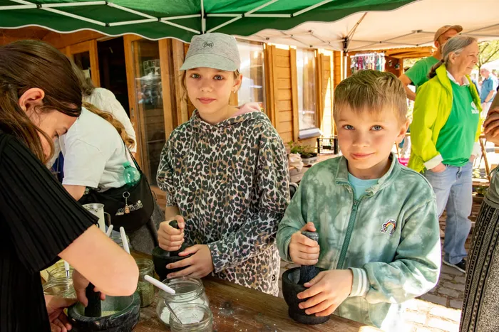Zwei Kinder stehen an einem Tisch und bearbeiten mit Mörsern Blüten. Im Hintergrund sind weitere Besucher und ein grüner Pavillon zu sehen.