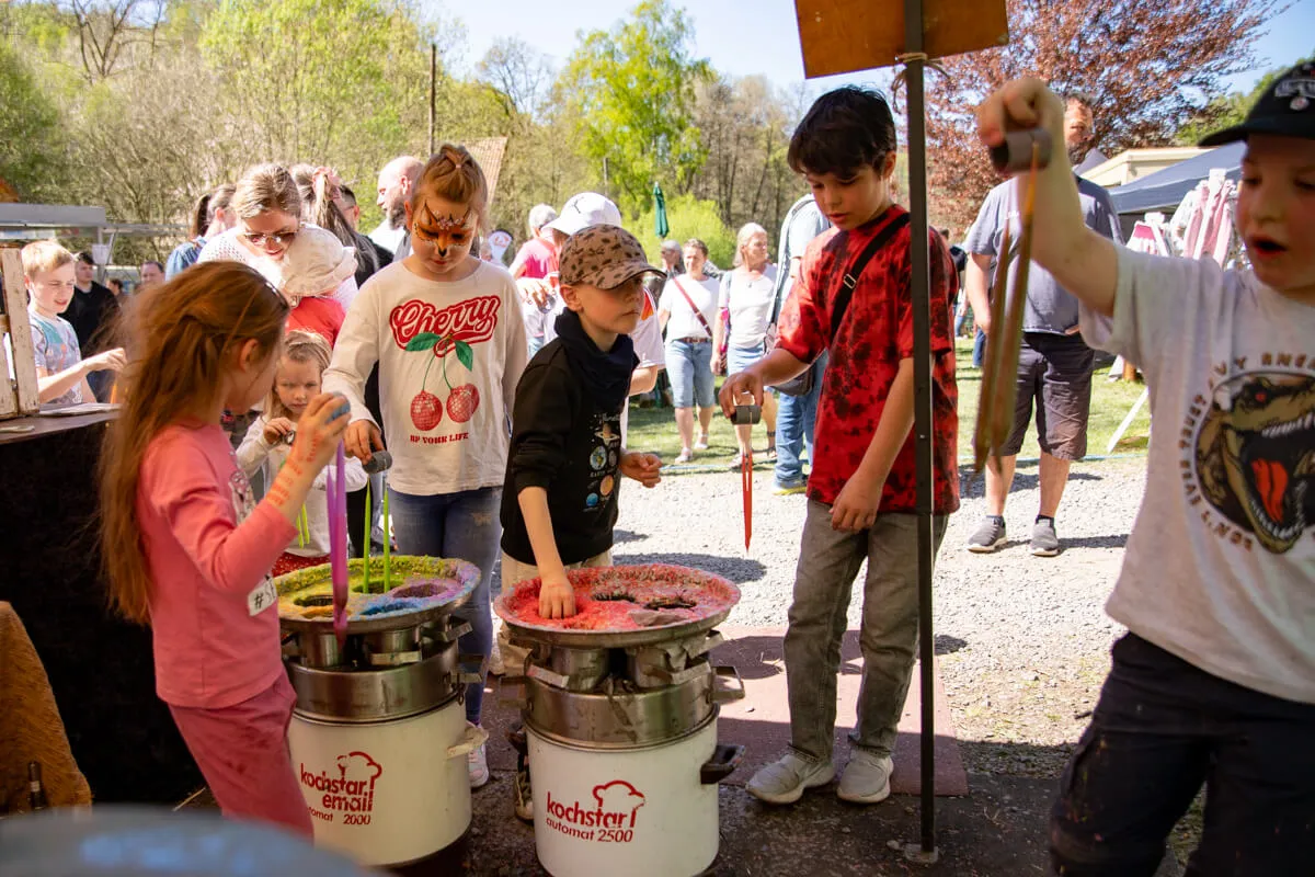 Kinder rühren mit langen Löffeln in großen Bottichen mit bunter Flüssigkeit, während andere zusehen. Im Hintergrund sind weitere Besucher und Zelte auf dem Gelände der Heesfelder Mühle zu erkennen.