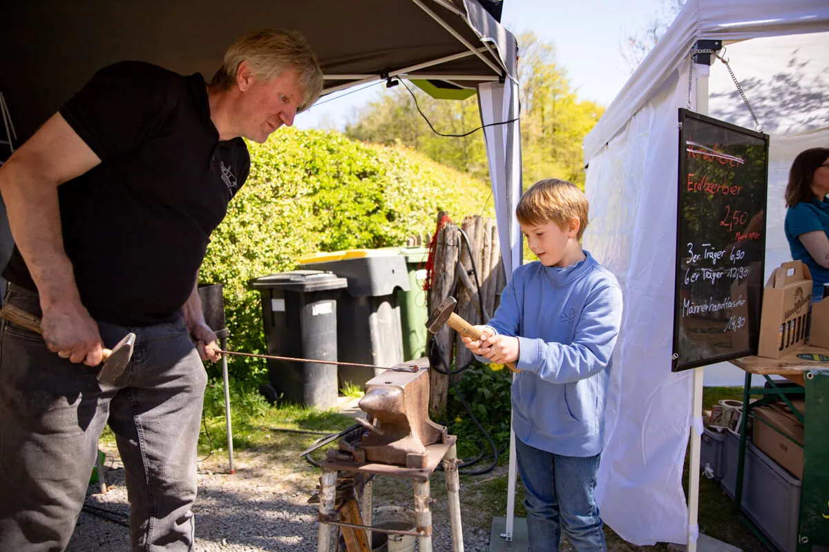 Ein Mann erklärt einem Jungen das Schmieden an einem mobilen Schmiedestand auf einem Frühlingsfest.