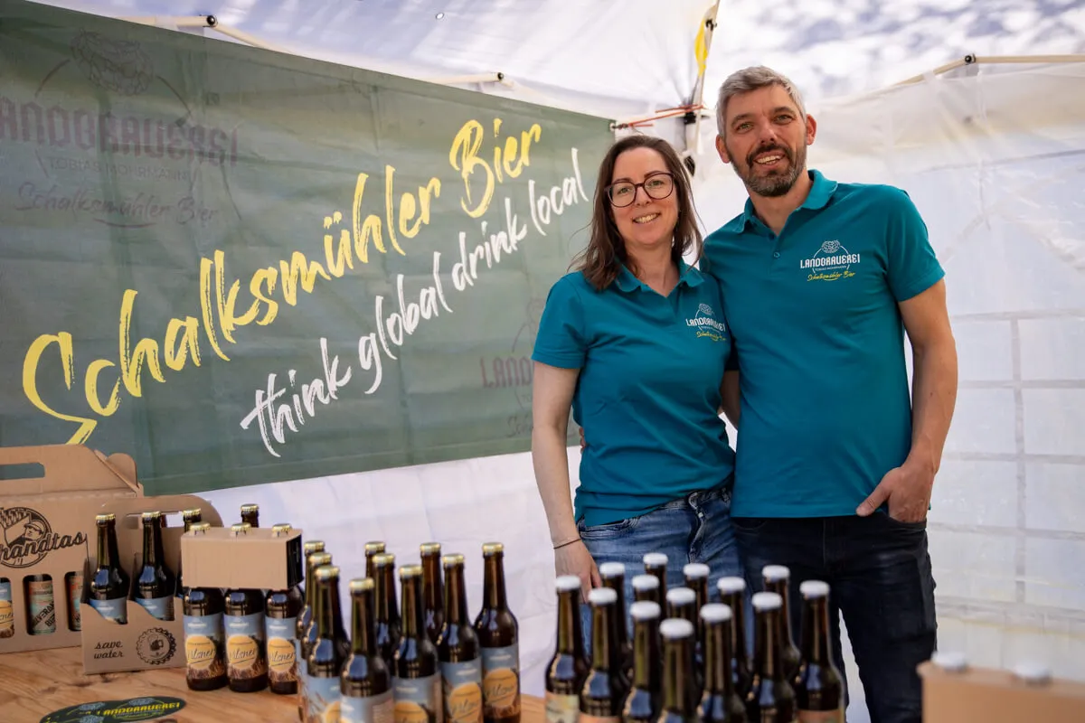 Zwei Menschen stehen lächelnd vor einem Stand mit Schalksmühler Bierflaschen und einem Banner mit der Aufschrift „Schalksmühler Bier think global drink local“.