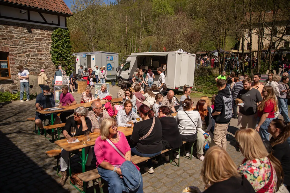 Menschen sitzen an Biertischgarnituren vor einer Fachwerkmühle und genießen Getränke und Gespräche bei einem Frühlingsfest.