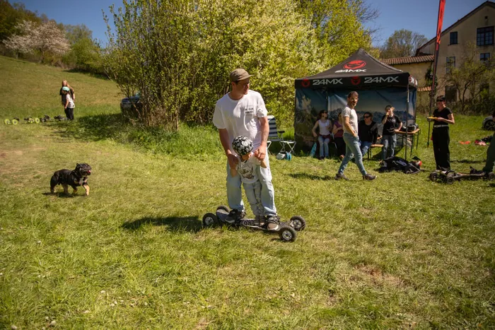 Ein Mann hilft einem Kind auf einem selbstgebauten Fahrzeug mit vier Rädern auf einer Wiese zu stehen. Im Hintergrund sind weitere Personen und ein Zelt mit der Aufschrift „24MX“ zu sehen.