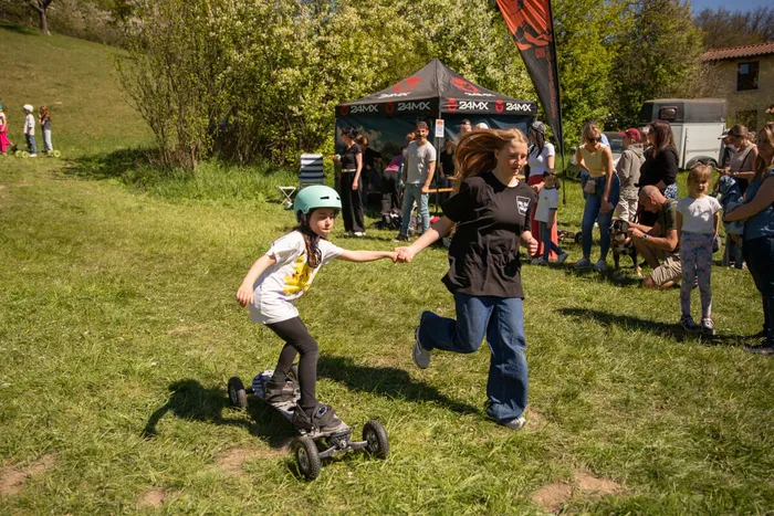 Ein Mädchen fährt auf einem Scooter mit vier Rädern, während eine junge Frau ihre Hand hält und mitläuft. Im Hintergrund stehen Menschen auf einer Wiese vor einem Zelt mit der Aufschrift „24MX“.