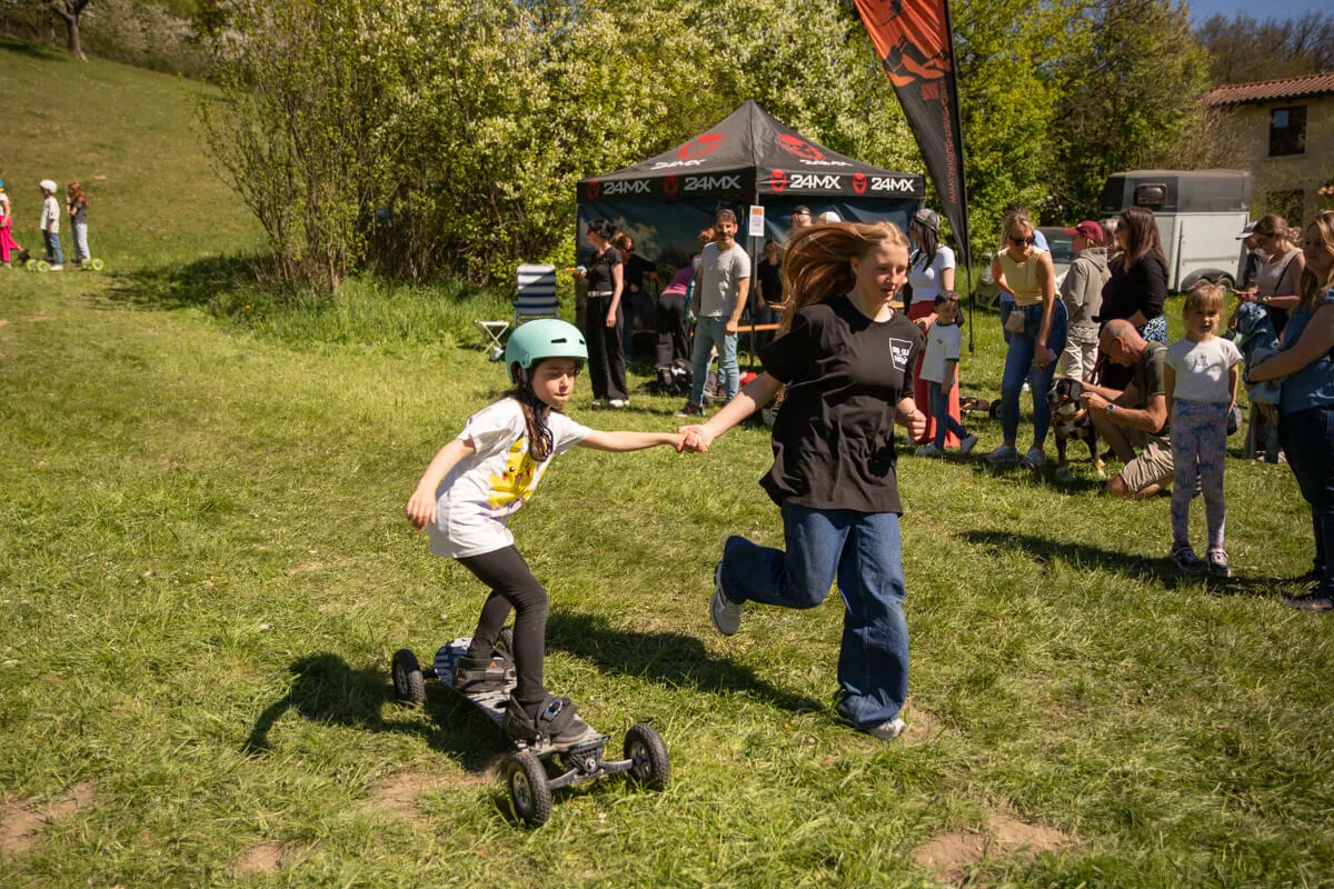 Ein Mädchen fährt auf einem Scooter mit vier Rädern, während eine junge Frau ihre Hand hält und mitläuft. Im Hintergrund stehen Menschen auf einer Wiese vor einem Zelt mit der Aufschrift „24MX“.