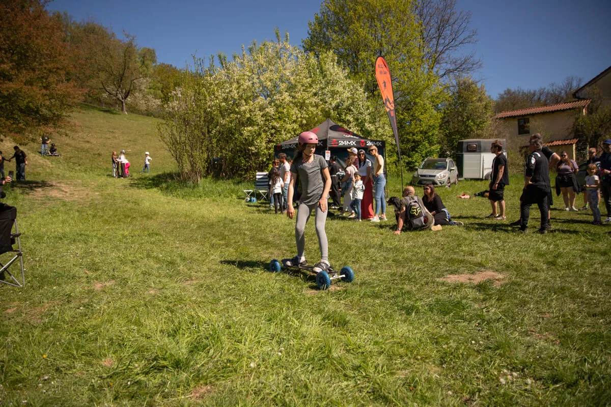 Eine Person fährt auf einem Longboard über eine Wiese, umgeben von Zuschauern und mehreren kleinen Zelten. Im Hintergrund ein Gebäude und blühende Bäume.
