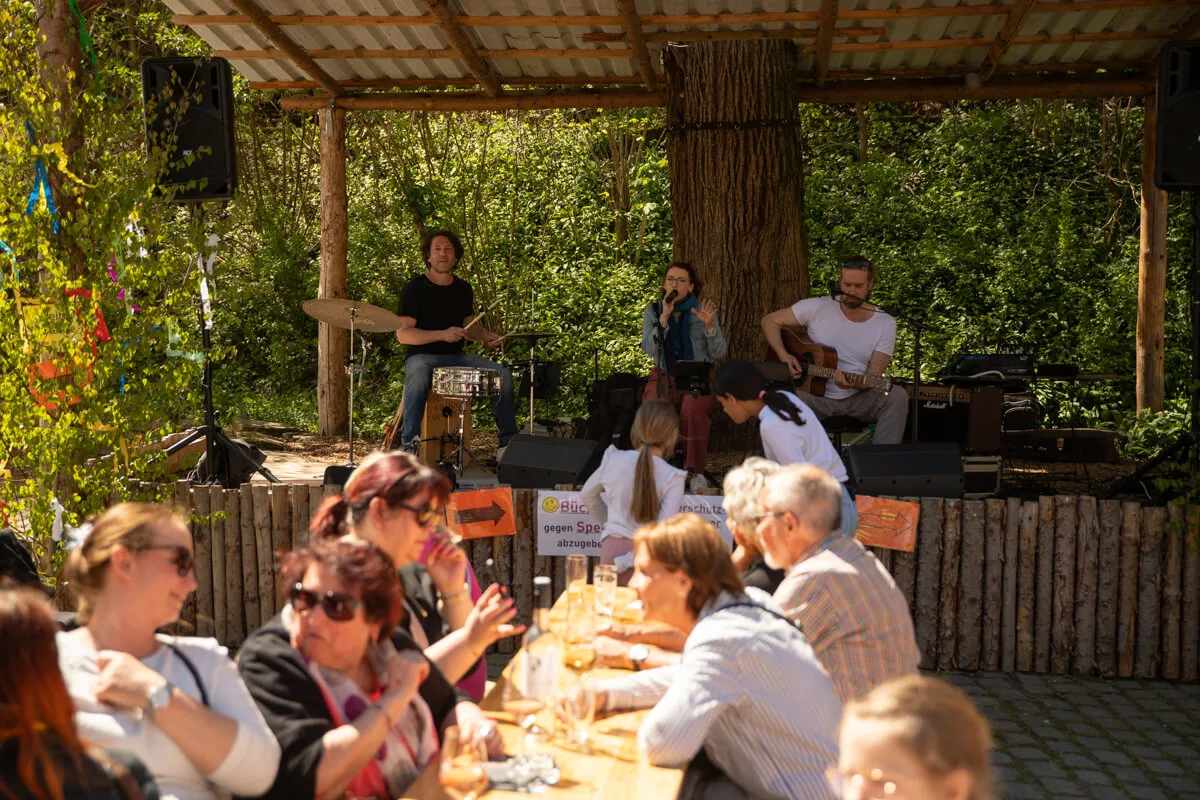 Drei Musiker spielen auf einer kleinen Bühne im Freien vor einem Publikum, das an Tischen sitzt. Im Hintergrund ein mit Pflanzen bewachsener Holzpavillon.
