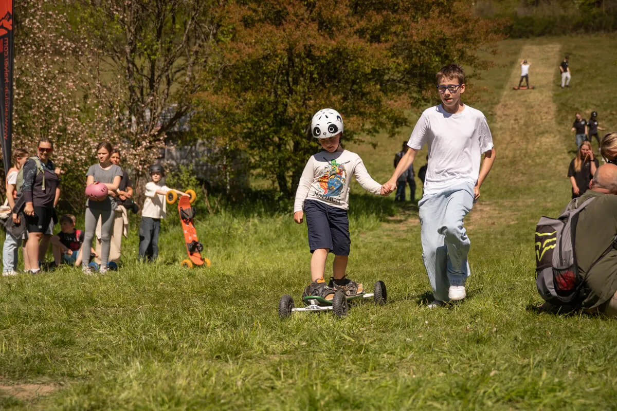 Ein Kind mit Helm fährt auf einem Roller, während ein Jugendlicher es an der Hand führt. Im Hintergrund sind weitere Menschen und Bäume zu sehen.