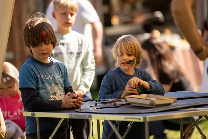 Zwei Jungen mit Gesichtsbemalung sitzen an einem Tisch im Freien und basteln. Im Hintergrund sind weitere Menschen und ein sonniger Tag zu sehen.