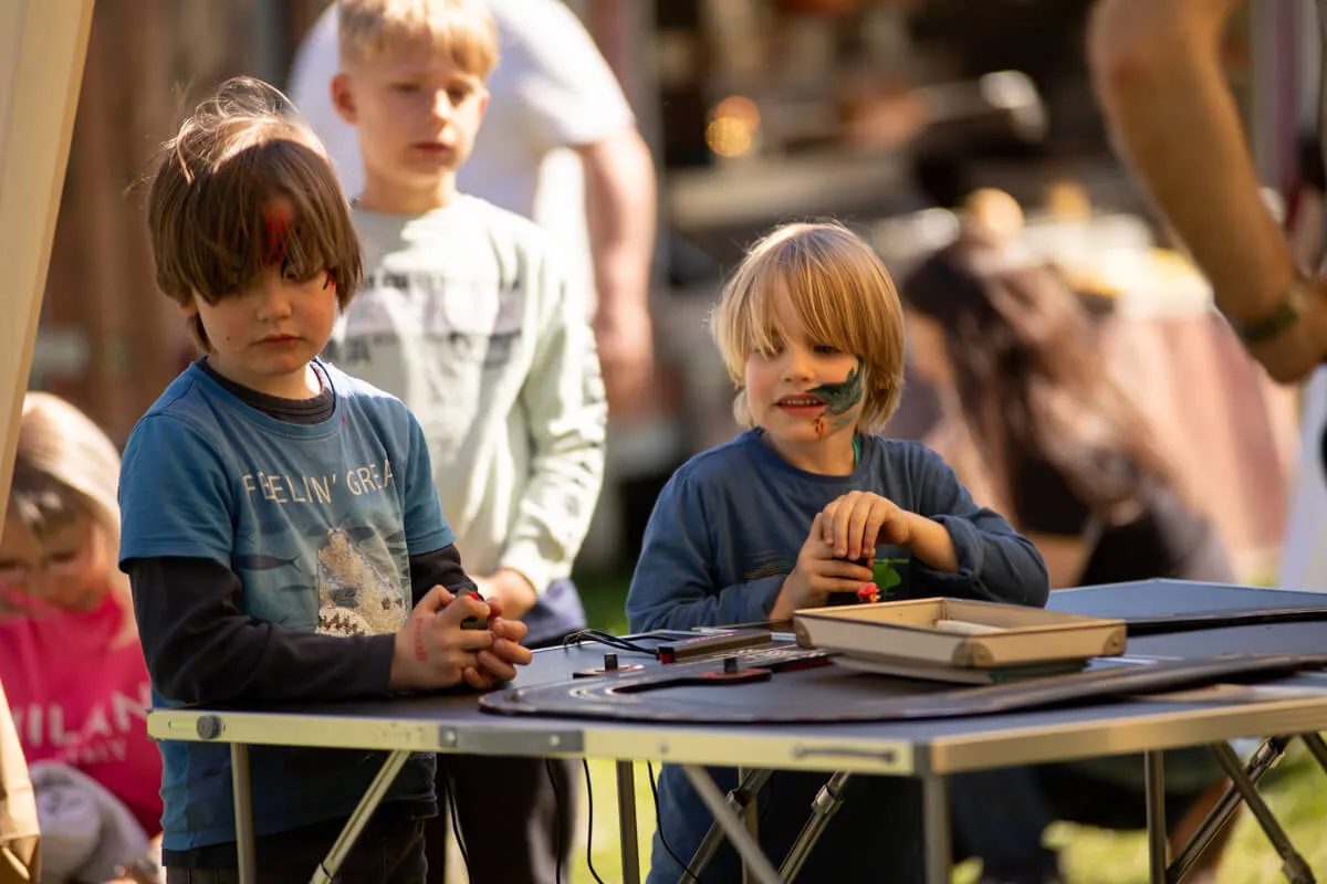 Zwei Jungen mit Gesichtsbemalung sitzen an einem Tisch im Freien und basteln. Im Hintergrund sind weitere Menschen und ein sonniger Tag zu sehen.