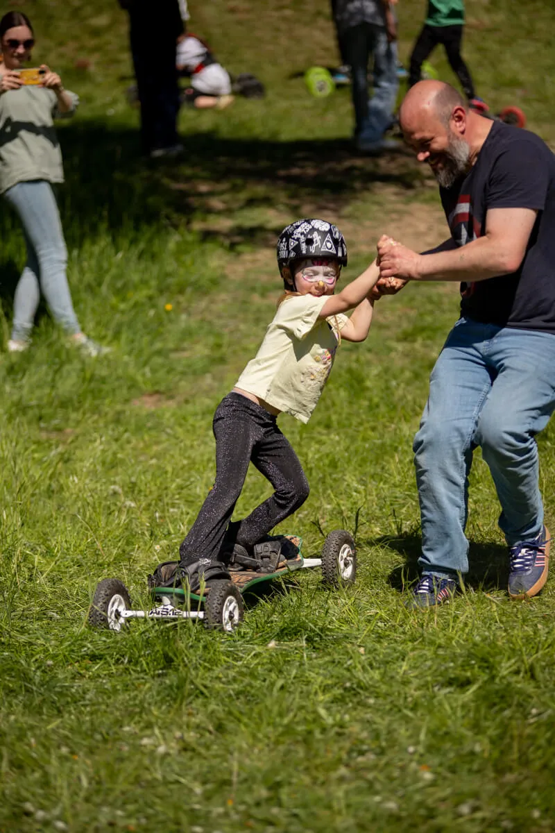 Ein Mann hält die Hand eines Kindes, das auf einem Rollbrett mit vier Rädern auf einer Wiese steht. Im Hintergrund sind weitere Menschen und ein Picknick zu sehen.