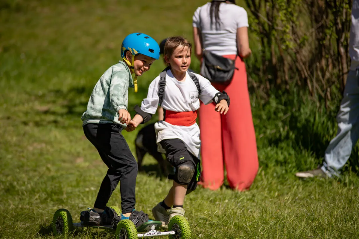 Zwei Kinder fahren auf einem selbstgebauten Gefährt mit Rollen über eine Wiese, wobei eines das andere schiebt. Im Hintergrund sind weitere Personen und Büsche zu sehen.