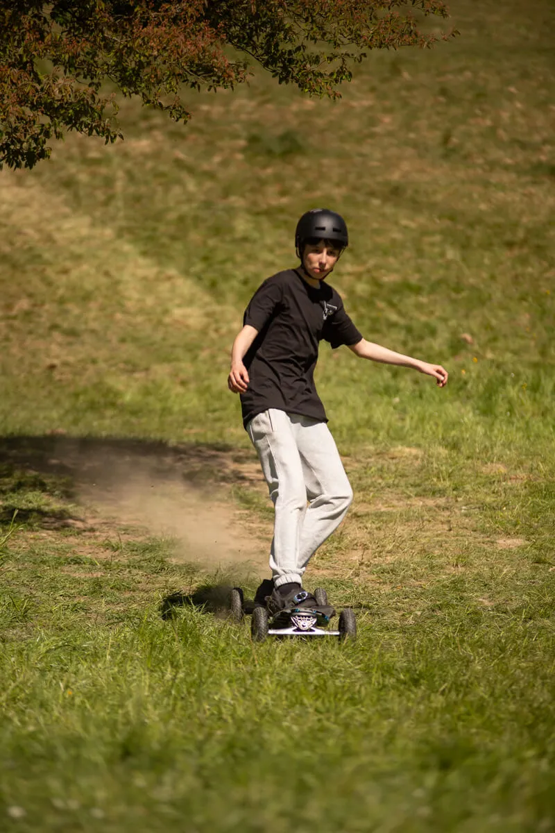 Ein Jugendlicher fährt mit einem elektrischen Skateboard auf einer Wiese, trägt einen Helm und lockere Kleidung.