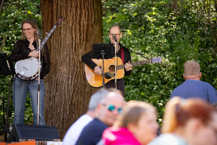 Zwei Musiker spielen auf einer kleinen Bühne im Freien: eine Frau mit einer Trommel und ein Mann mit einer Gitarre. Im Hintergrund sitzen und stehen Besucher.