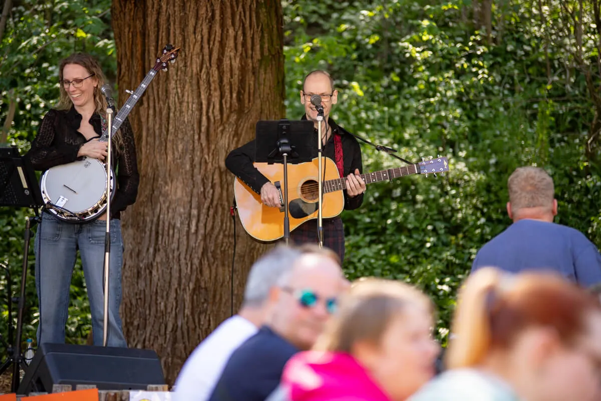 Zwei Musiker spielen auf einer kleinen Bühne im Freien: eine Frau mit einer Trommel und ein Mann mit einer Gitarre. Im Hintergrund sitzen und stehen Besucher.
