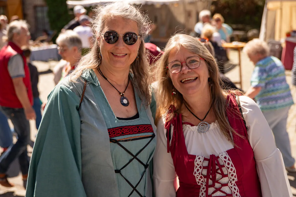 Zwei lächelnde Frauen mit langen, lockigen Haaren tragen mittelalterliche Kleidung und Schmuck auf dem Kirschblütenfest an der Heesfelder Mühle.