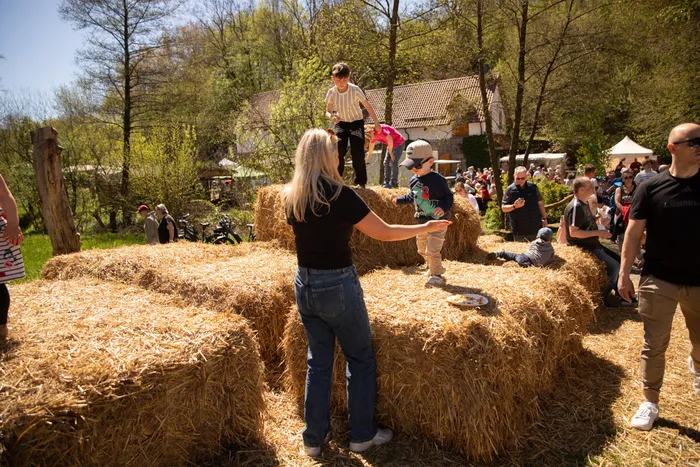 Eine Frau steht vor Heuballen, während Kinder darauf klettern. Im Hintergrund sind weitere Menschen und Gebäude auf dem Gelände der Heesfelder Mühle zu sehen.