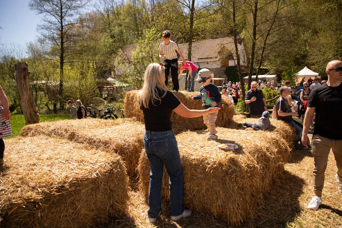 Eine Frau steht vor Heuballen, während Kinder darauf klettern. Im Hintergrund sind weitere Menschen und Gebäude auf dem Gelände der Heesfelder Mühle zu sehen.
