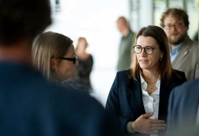 Ina Blumenthal, eine Frau mit Brille und Blazer, spricht auf einem Gipfel mit einer anderen Frau. Im Hintergrund sind weitere Personen zu sehen.