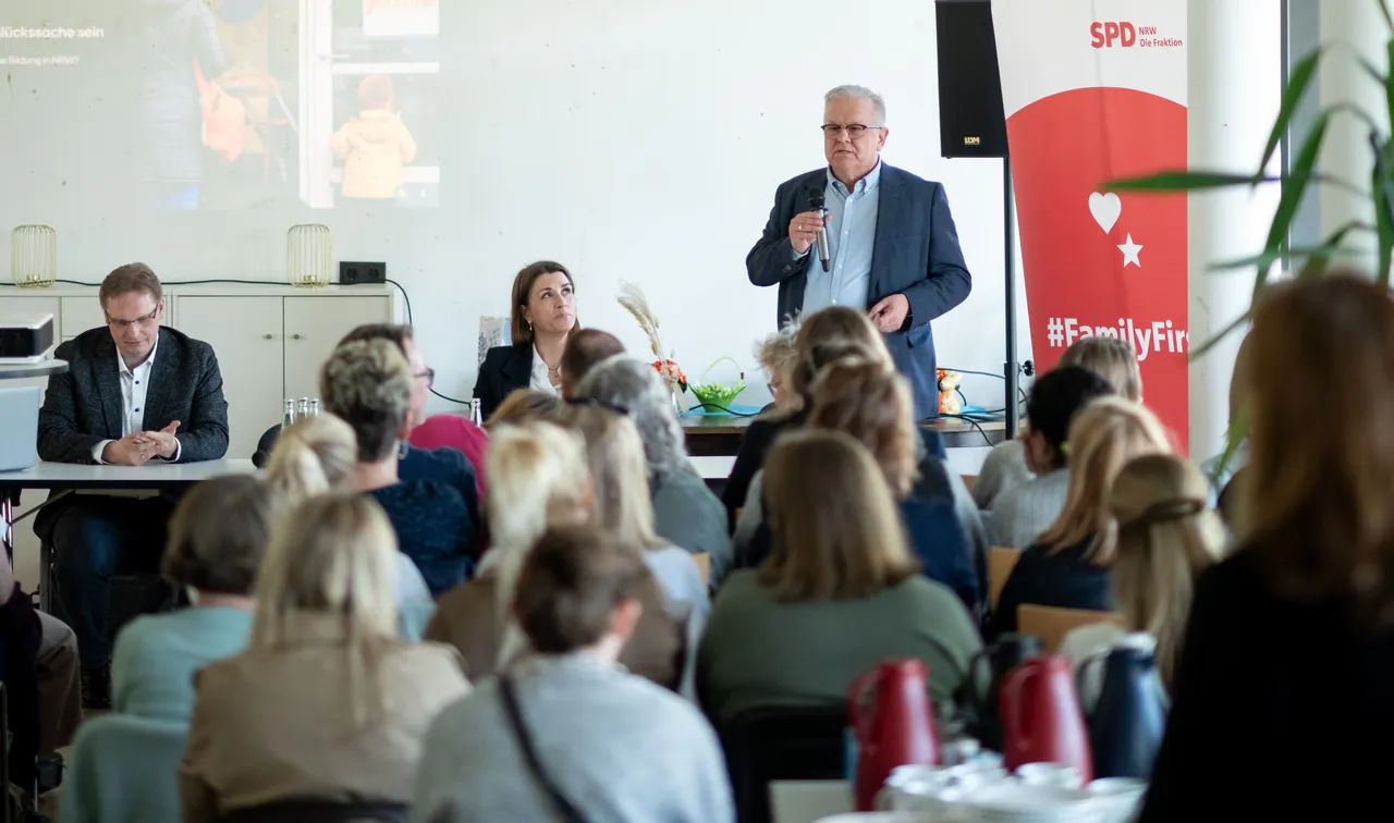 Wolfgang Jörg spricht auf einem Kita-Gipfel vor einem Publikum, während zwei weitere Personen am Tisch sitzen und zuhören. Im Hintergrund ist ein Bildschirm mit einer Präsentation zu sehen.