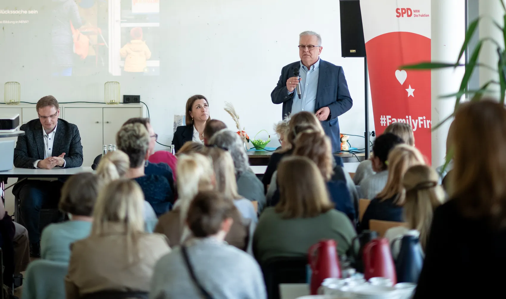 Wolfgang Jörg spricht auf einem Kita-Gipfel vor einem Publikum, während zwei weitere Personen am Tisch sitzen und zuhören. Im Hintergrund ist ein Bildschirm mit einer Präsentation zu sehen.
