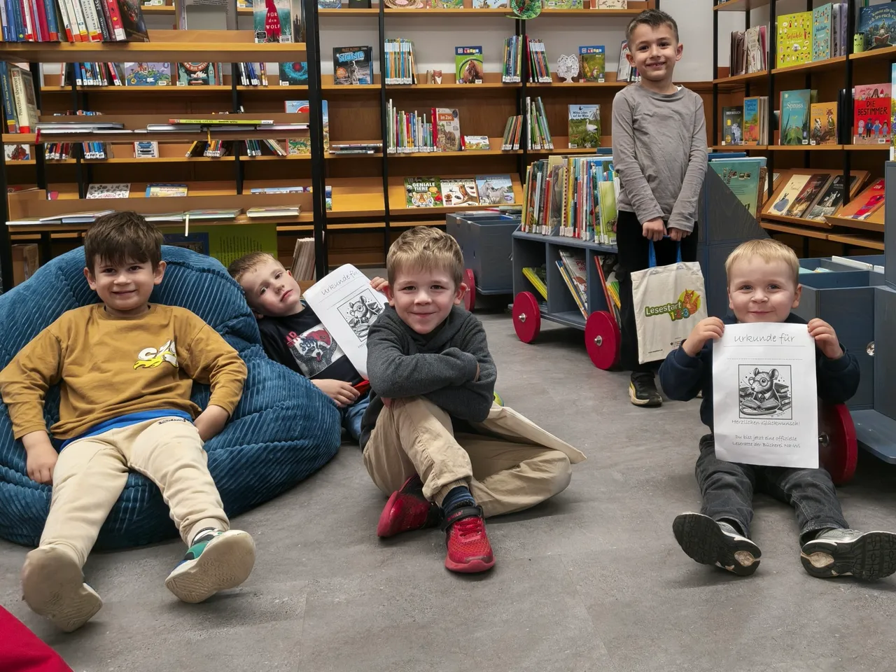 Fünf Kinder sitzen oder lehnen in einer Bibliothek, einige halten Urkunden mit einem Tiermotiv. Im Hintergrund stehen Regale voller Bücher.