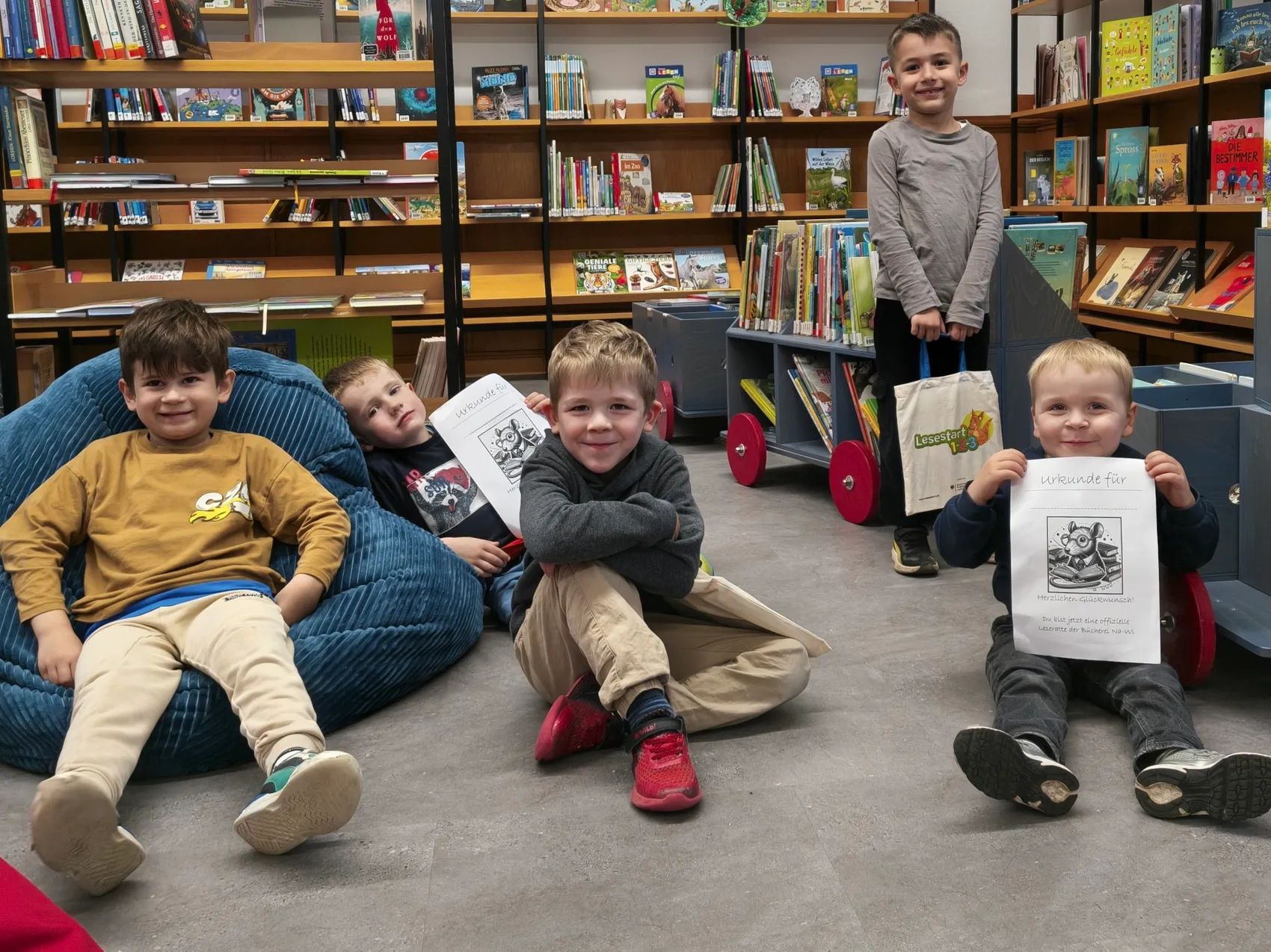 Fünf Kinder sitzen oder lehnen in einer Bibliothek, einige halten Urkunden mit einem Tiermotiv. Im Hintergrund stehen Regale voller Bücher.