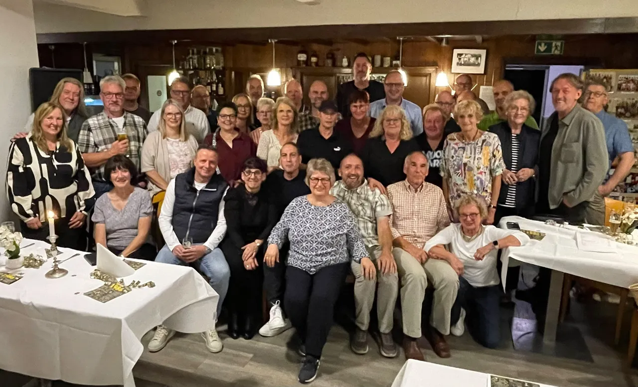 Eine große Gruppe ehemaliger Klassenkameraden posiert für ein Gruppenfoto bei einem Klassentreffen an weiß gedeckten Tischen.