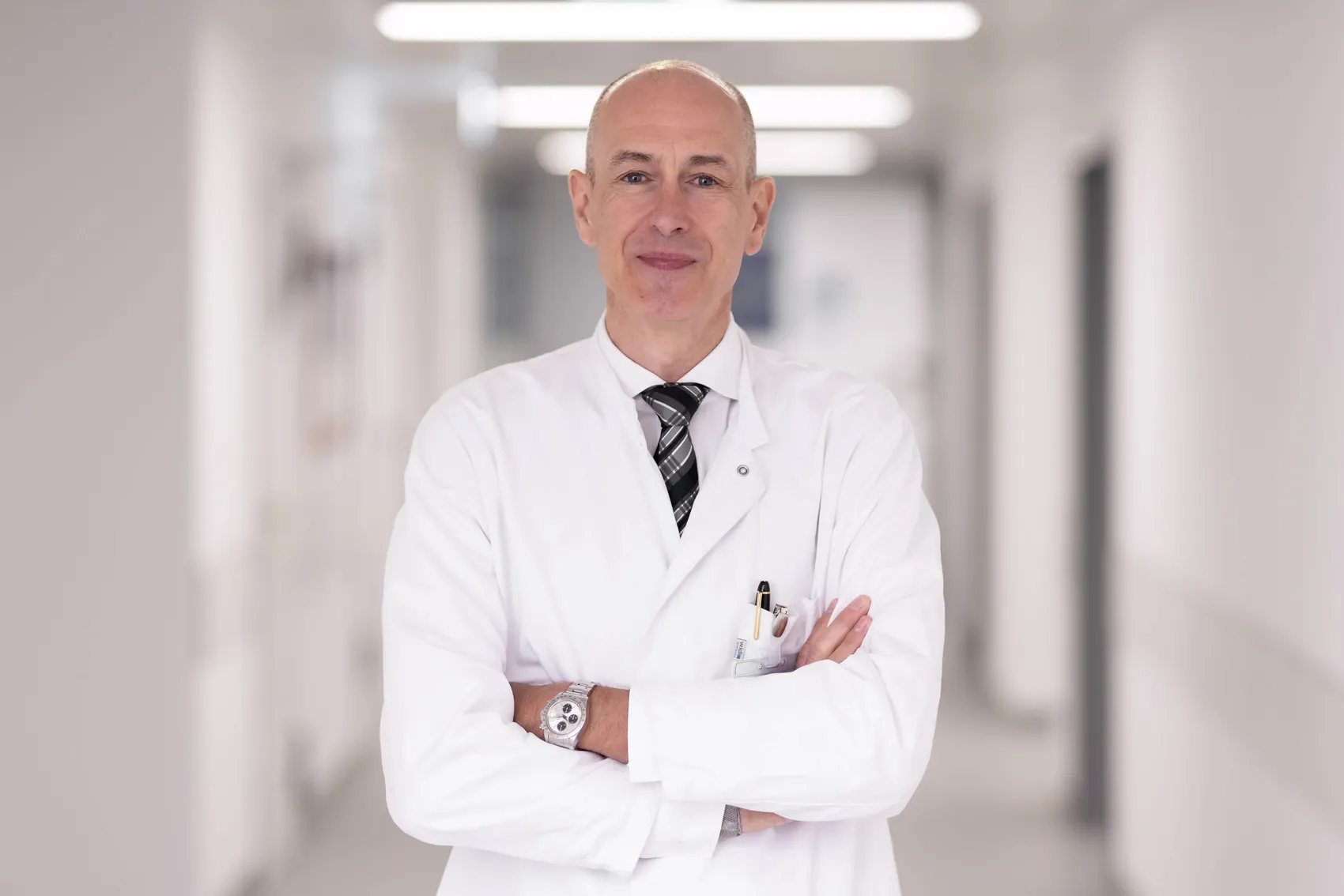 Frank Beckers in weißem Arztkittel und Krawatte mit verschränkten Armen in einem hellen Krankenhausflur.