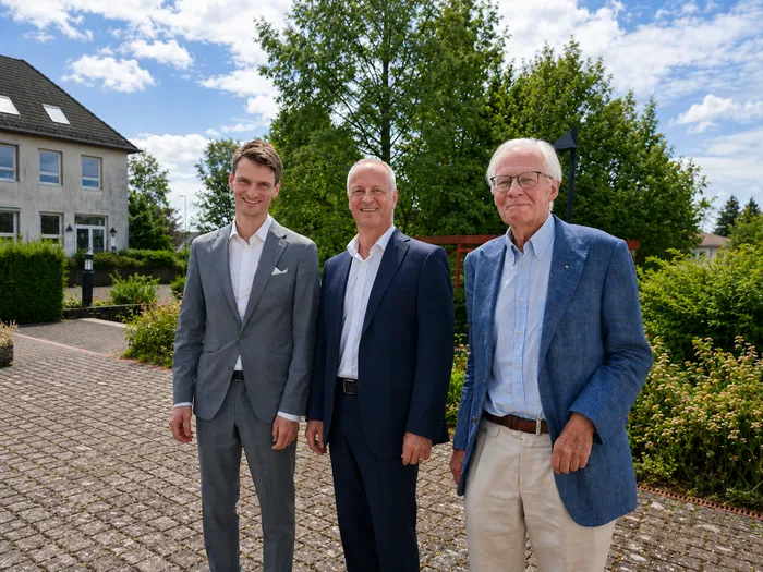 Drei Männer in formeller Kleidung stehen nebeneinander vor einem Gebäude und Hecken. Der jüngste trägt einen hellgrauen Anzug, der mittlere einen dunkelblauen und der älteste einen blauen Blazer zu beigen Hosen. Das Bild zeigt den Führungswechsel der Nephrologie am Klinikum Lüdenscheid.