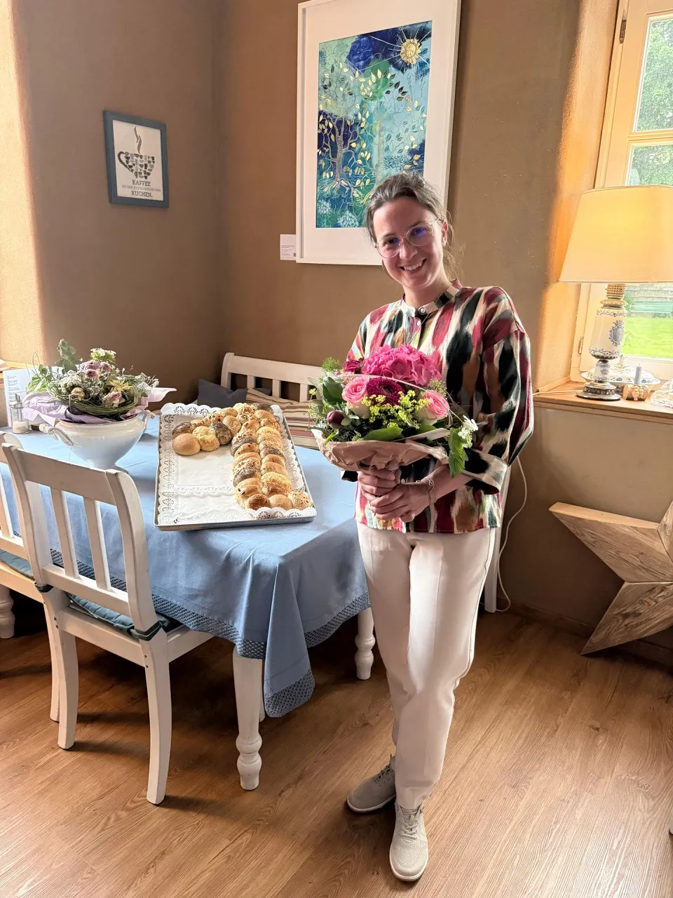 Frau mit Blumenstrauß und Gebäck auf einem Tisch im Café Klön. Im Hintergrund ein Bild mit stilisierter Sonnenblume.
