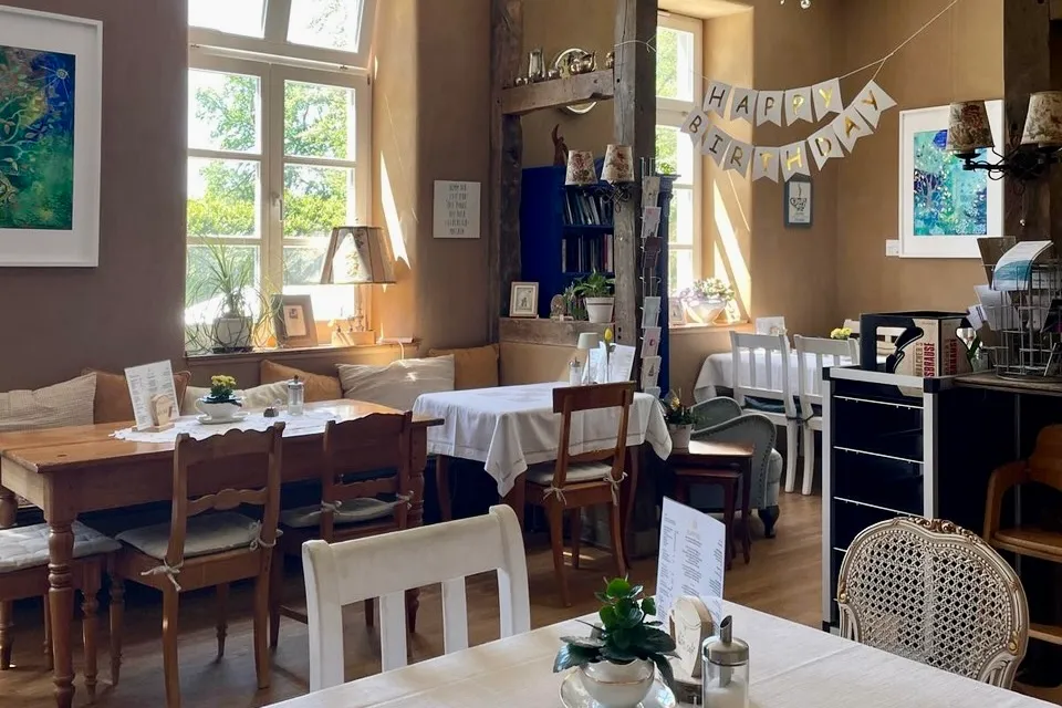 Innenansicht des Klön Cafés mit Tischen und Stühlen, großen Fenstern und einer Girlande mit der Aufschrift „Happy Birthday“.