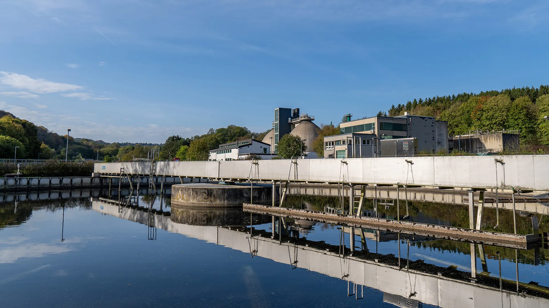 Die Kläranlage Kohlfurth mit ihren Gebäuden und Becken, die sich im Wasser spiegeln, vor bewaldeter Hügellandschaft.