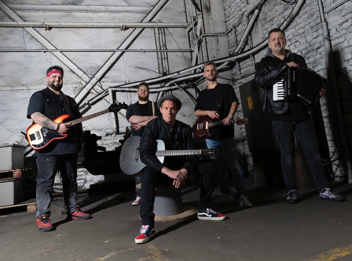 Fünf Musiker posieren in einem industriell wirkenden Raum mit freiliegenden Rohren und Ziegelwänden. Sie spielen verschiedene Instrumente wie Gitarre, Bass und Akkordeon.