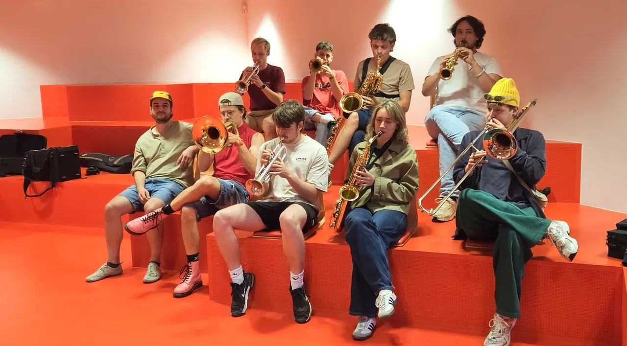 Die Mitglieder der Brassband Knallblech sitzen auf roten Stufen und spielen gemeinsam verschiedene Blasinstrumente wie Trompeten, Posaunen und Saxophone.