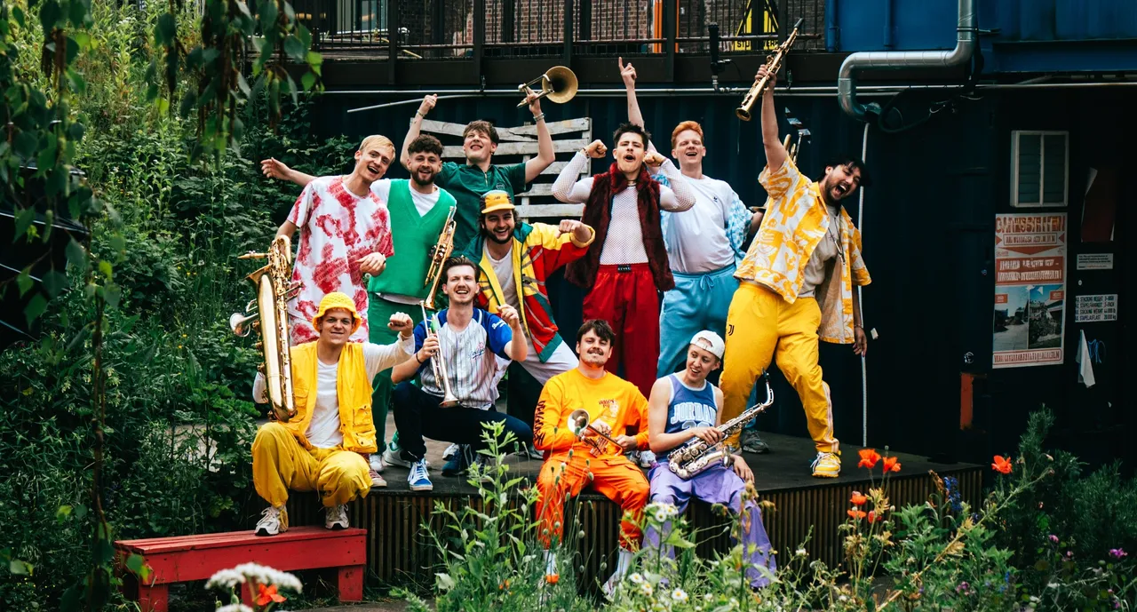 Die Musikgruppe „Knallblech“ posiert in farbenfrohen Outfits mit Blasinstrumenten vor einer Kulisse aus Pflanzen und einem blauen Container.