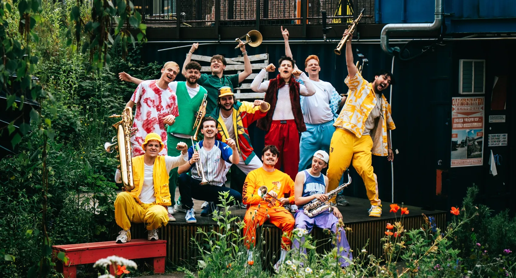 Die Musikgruppe „Knallblech“ posiert in farbenfrohen Outfits mit Blasinstrumenten vor einer Kulisse aus Pflanzen und einem blauen Container.
