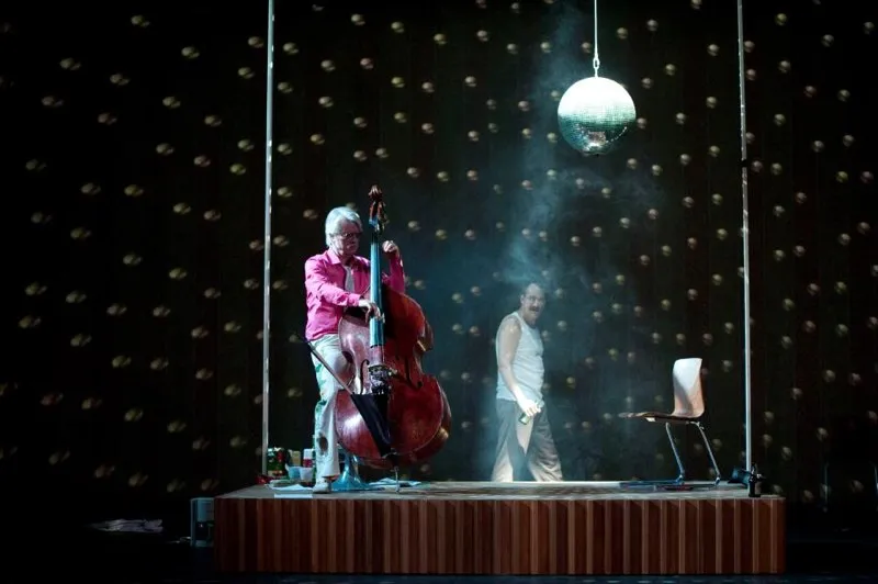 Ein Mann spielt auf einem Kontrabass auf einer erhöhten Bühne, während ein anderer Mann im Hintergrund einen Stuhl hält. Über der Bühne hängt eine Discokugel.
