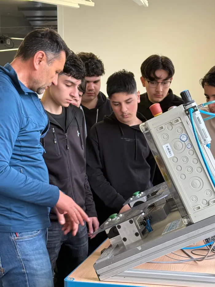 Ein Mann zeigt Schülern an einer metallenen Maschine auf einen bestimmten Punkt. Die Schüler beobachten aufmerksam.