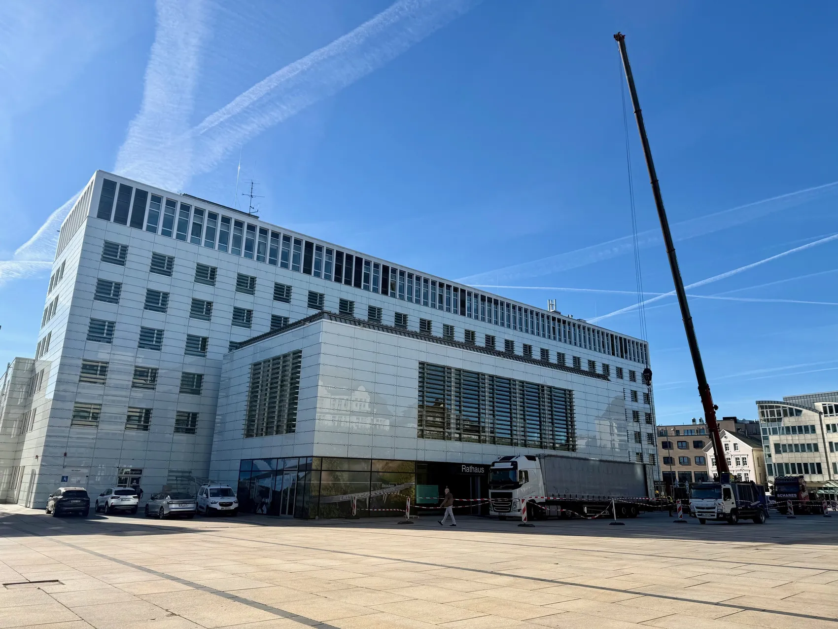 Ein Kran hebt eine Klimaanlage auf das Dach eines modernen, weißen Rathauses auf einem belebten Platz.