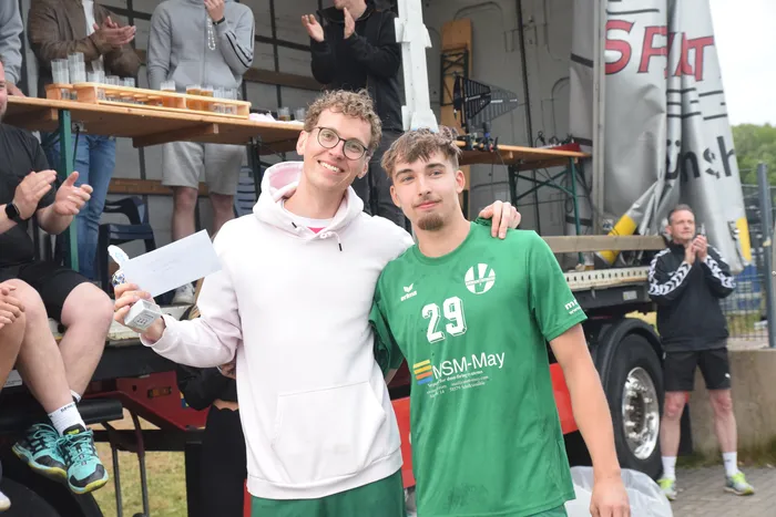 Zwei junge Männer umarmen sich vor einer Menschenmenge und einem Getränkestand. Der Mann rechts trägt ein Handballtrikot mit der Nummer 29.