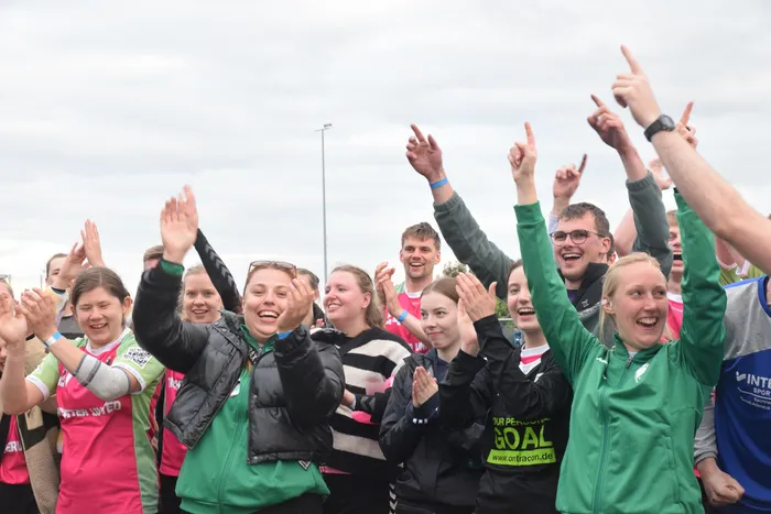 Jubelnde Menschen mit hoch erhobenen Armen stehen auf einem Sportplatz. Viele tragen Sportkleidung in den Farben Grün, Pink und Schwarz.