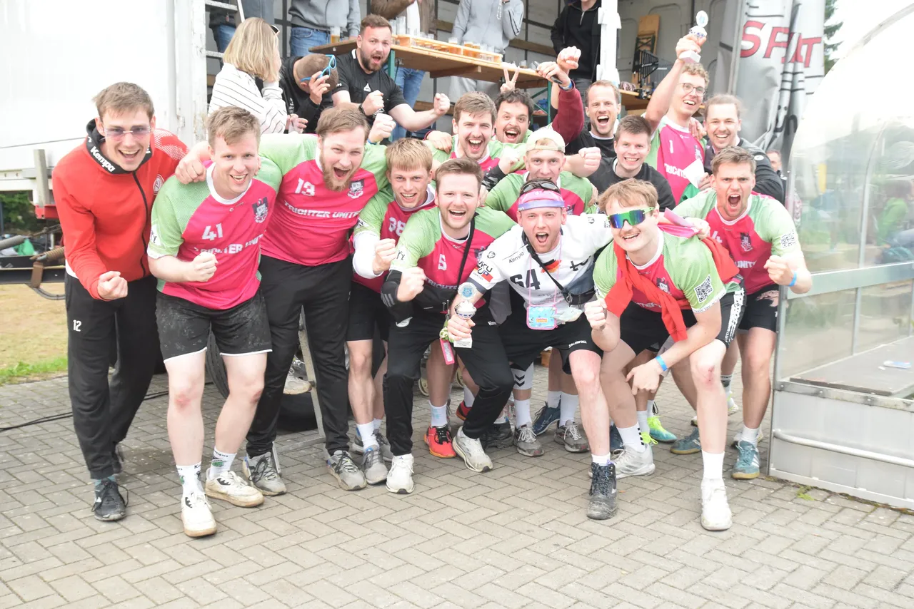 Eine Gruppe junger Männer und Frauen in Handballtrikots feiert ausgelassen und jubelt in die Kamera. Einige halten Becher oder Medaillen hoch. Im Hintergrund sind weitere Personen und ein Imbisswagen zu sehen.